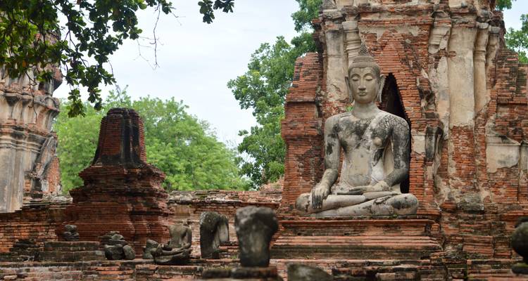 Statue ancienne de Bouddha assis parmi les ruines en briques rouges qui s'effritent et les arbres feuillus sur un site de temple thaï historique.