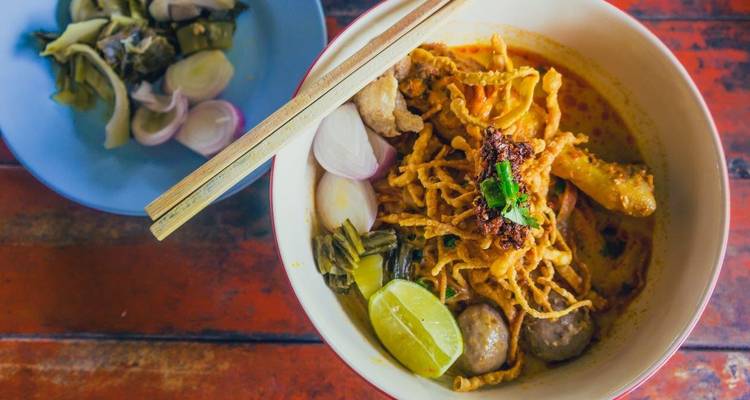 Un bol de soupe de nouilles Khao Soi crémeuse garni de citron vert, de nouilles frites et d'herbes sur une table en bois rustique.