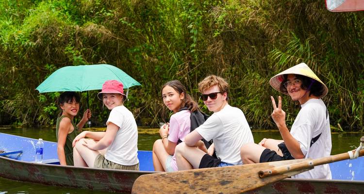 Jóvenes viajeros alegres se sientan en un bote estrecho en un río de la selva, algunos sosteniendo paraguas y haciendo señales de paz.