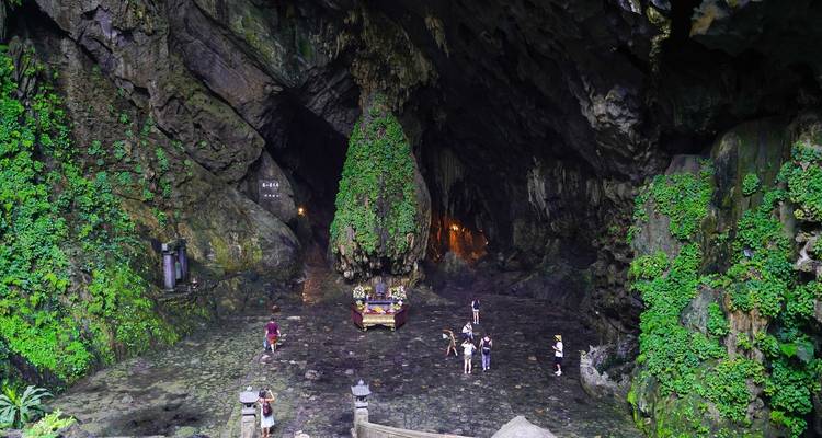 Vasta cámara de cueva cubierta de verde alberga un pequeño santuario, con visitantes explorando el suelo rocoso.