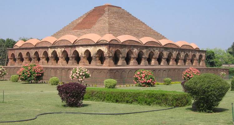 Pyramidal Rasmancha temple with arch colonnades surrounded by blooming shrubs under clear sky.