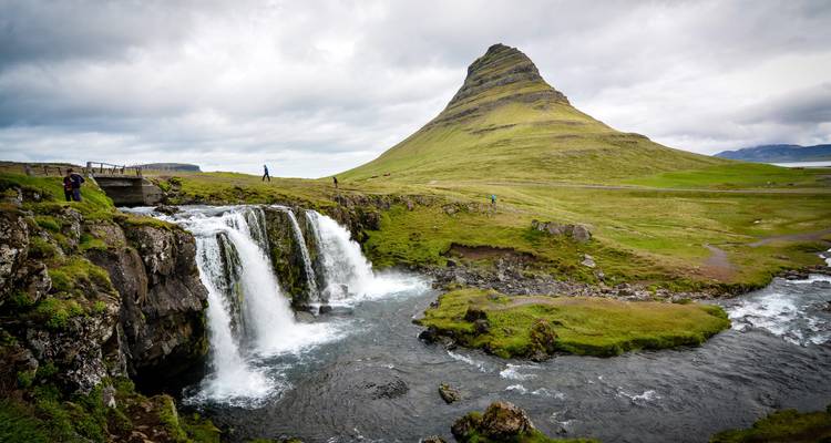La montagne Kirkjufell s'élève derrière une cascade jumelle qui dévale à travers des falaises vertes et mousseuses lors d'une journée couverte.