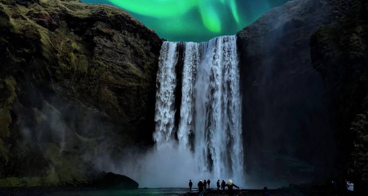 Des visiteurs en silhouette se tiennent au pied de la cascade de Skogafoss alors que des aurores vertes ondulent dans le ciel nocturne au-dessus.