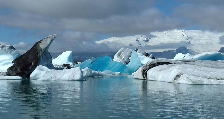 Des icebergs aux formes irrégulières, bleus et blancs, flottent dans les eaux calmes du lagon de Jökulsárlón sous des pics enneigés.