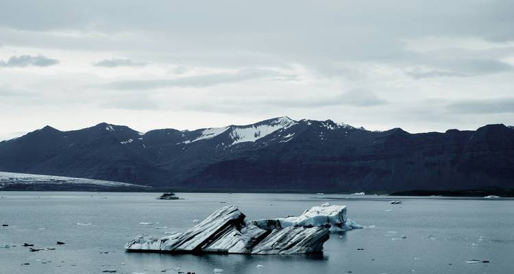 Un iceberg solitaire aux stries distinctes dérive dans un lagon glaciaire gris encadré par des montagnes sombres et escarpées.
