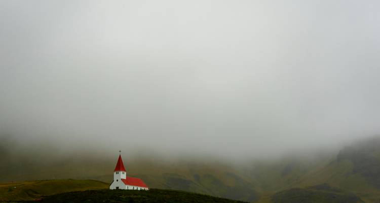 A small white church with a red roof sits beneath a blanket of low fog on a grassy Icelandic hill.
