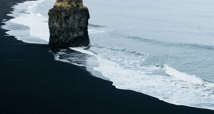 Waves lap against a black-sand beach where a solitary sea stack rises from the North Atlantic.