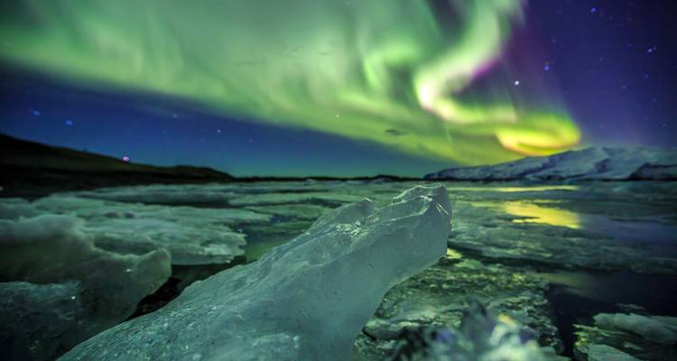 La aurora boreal de un verde brillante baila sobre bloques de hielo glacial y una laguna reflectante bajo cielos estrellados.