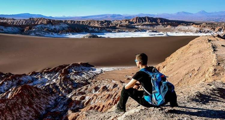 Ein Reisender mit Rucksack sitzt auf einem Wüstenkamm und blickt auf die weiten, unwirklichen Dünen und Täler der Atacama.