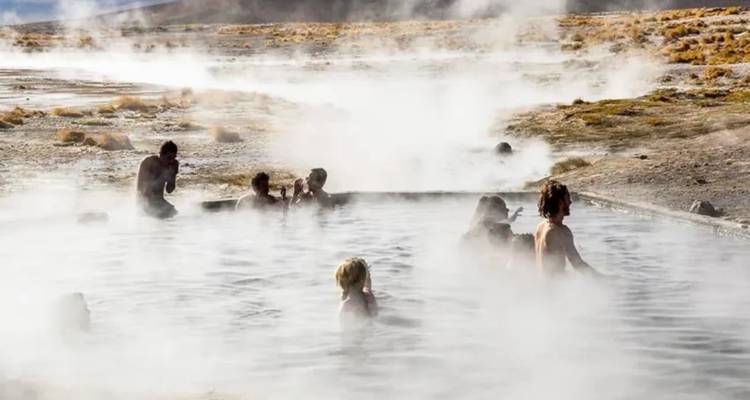 Mehrere Reisende baden in einem dampfenden geothermischen Becken, das von trockener Landschaft umgeben ist.