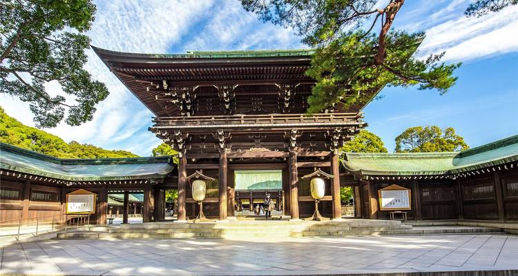 Traditionelles Tor im Torii-Stil aus Holz, das zu einem ruhigen Shinto-Schrein-Komplex führt, umgeben von blauem Himmel und Bäumen.