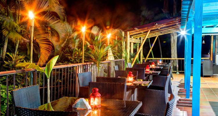Terraza de restaurante al aire libre iluminada por faroles y antorchas entre plantas tropicales exuberantes por la noche.