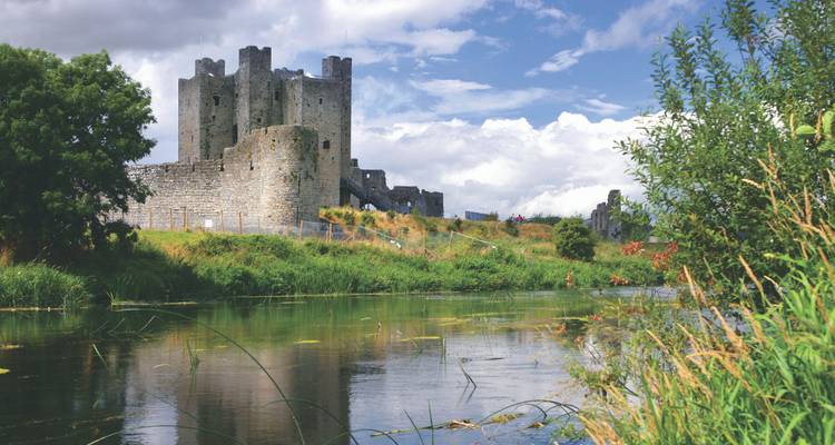 Château de pierre médiéval en bord de rivière se reflétant dans des eaux calmes sous un ciel partiellement nuageux