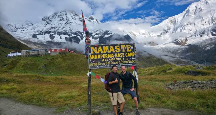 Two hikers posing by the Annapurna Base Camp welcome sign with dramatic snowy mountains