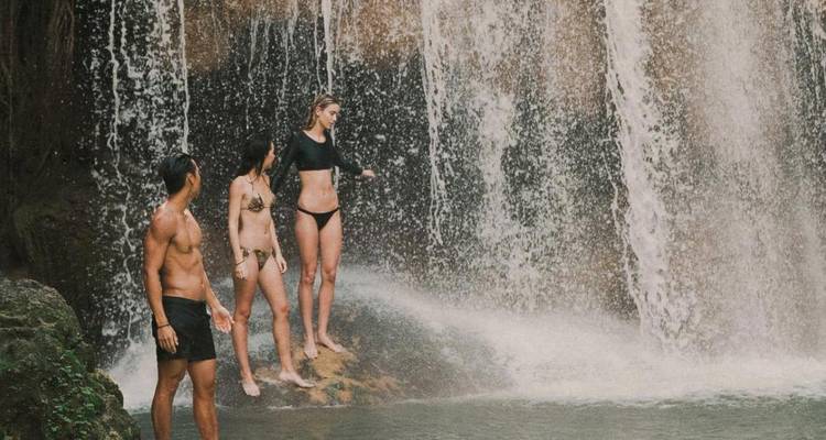 Three travelers in swimwear standing beneath a cascading tropical waterfall