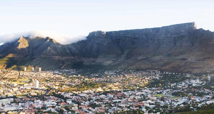 Panoramic daytime view of Cape Town with sprawling cityscape in the foreground and the dramatic cliffs of Table Mountain rising behind, partly shrouded by a cloud bank.