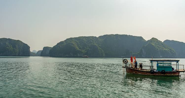 Kleines hölzernes Fischerboot mit vietnamesischen Flaggen gleitet über ruhiges smaragdgrünes Wasser, hinter dem sich imposante Kalkstein-Karstinseln erheben.