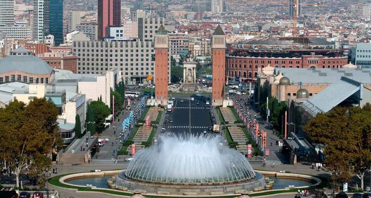 Amplio paisaje urbano de la Plaça d'Espanya de Barcelona con la Fuente Mágica, las torres venecianas gemelas y la antigua plaza de toros, bajo un cielo azul despejado.