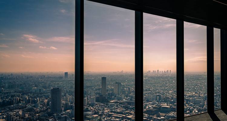 Hazy sunset cityscape of Tokyo viewed through tall glass windows of a skyscraper observation deck.