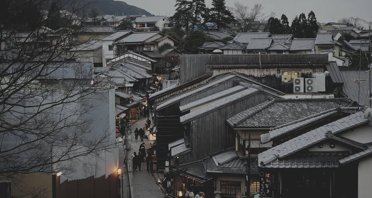 Narrow historic Japanese street lined with wooden houses and evening lights, bustling with pedestrians.