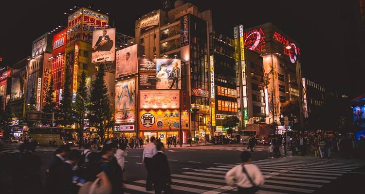 Vibrant neon-lit Tokyo street crossing jammed with people beneath towering advertising screens at night.