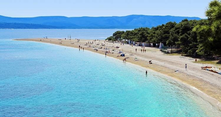 Een prachtig strand met helder turquoise water en mensen die ontspannen op de zandige kust, met dennenbomen en bergen op de achtergrond.