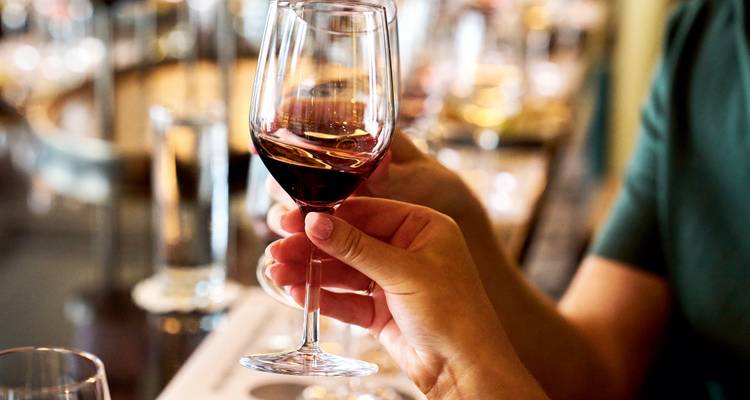 Gros plan d'une main qui fait tournoyer un verre de vin rouge lors d'une séance de dégustation intérieure, avec d'autres verres flous en arrière-plan.