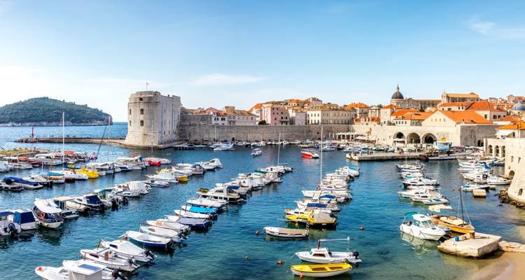 Vue panoramique du vieux port de Dubrovnik rempli de petits bateaux, encadré par des murs en pierre et des toits aux tuiles orange sous un ciel bleu dégagé.