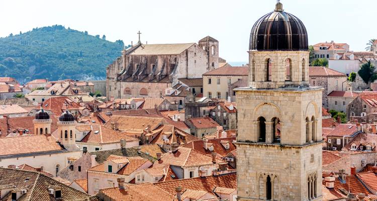 Vue élevée sur les toits en terre cuite de Dubrovnik avec un clocher prominent et des collines verdoyantes au loin.