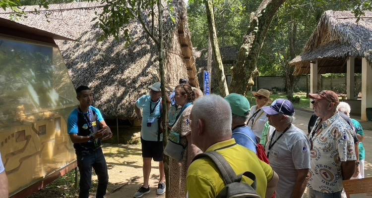 Tour group listening to a local guide beside a display board under thatched shelters in Cu Chi, Vietnam.