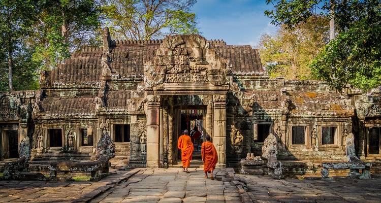 Two monks in orange robes walk toward the entrance of an ancient stone temple surrounded by trees.