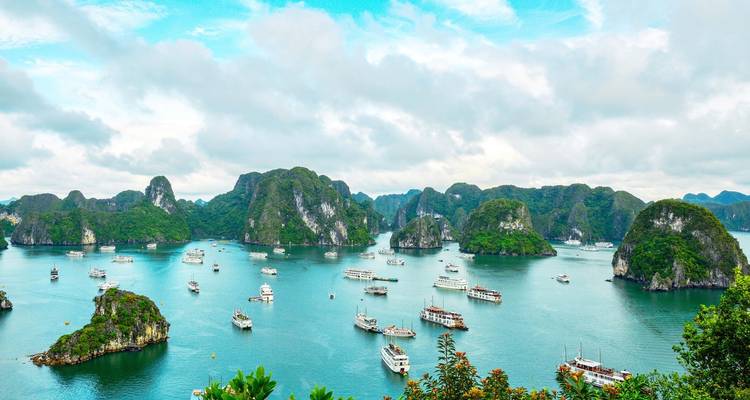 Wide aerial of Halong Bay with numerous cruise boats sailing amid forested limestone pillars and turquoise water.