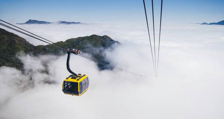 Bright yellow cable car glides above a sea of clouds toward rugged mountain peaks.
