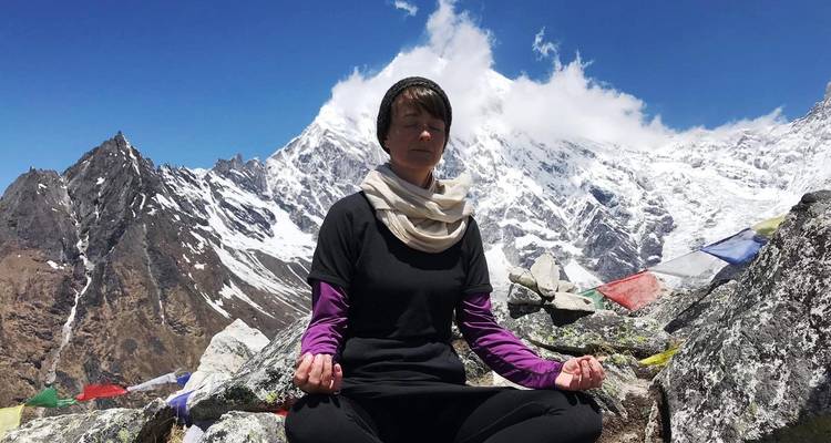 Persoon die mediteert op rotsachtig terrein met gebedsvlaggen en besneeuwde bergen.