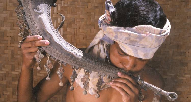 Man in traditional headwear playing a tribal wind instrument.