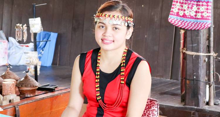 Smiling young woman in traditional Sabah attire with beaded necklace inside a wooden house.