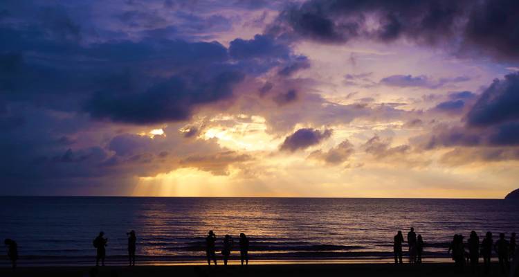 Silhouettes de personnes sur une plage tropicale regardant les rayons spectaculaires du coucher de soleil perçant à travers des nuages violets au-dessus de la mer.