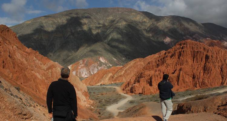 Zwei Menschen betrachten bunte Gesteinsformationen vor einer Bergkulisse.