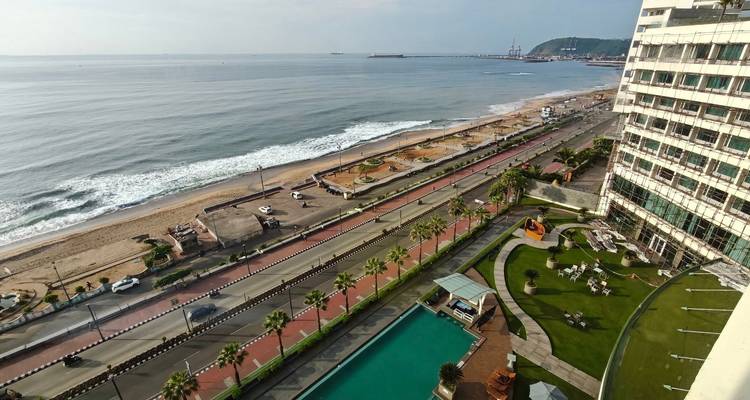 Vue en plongée d'un boulevard côtier bordé de palmiers, d'un front de mer, de vagues douces et d'une terrasse de piscine d'hôtel.