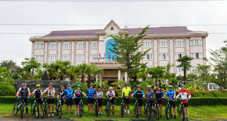 Des cyclistes se tiennent debout avec leurs vélos devant la façade d'un hôtel pour un portrait de groupe par une matinée nuageuse.