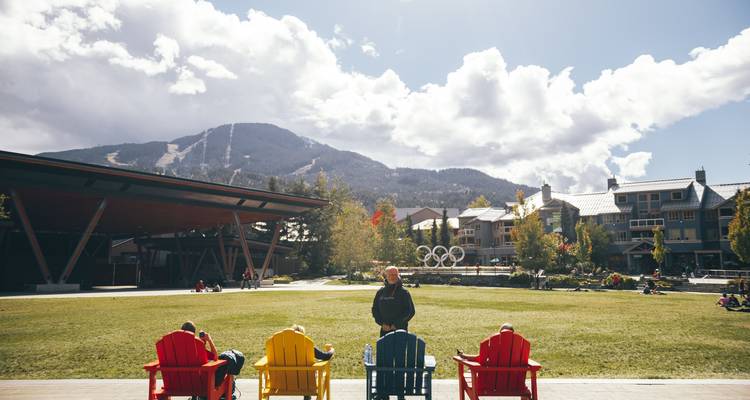 Open grassy plaza with colorful Adirondack chairs facing ski slopes and alpine village buildings under a bright sky.