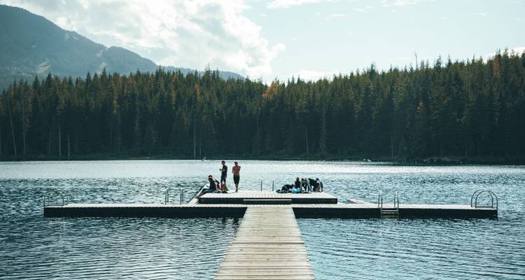 A wooden dock jutting into a forested lake where small groups gather to enjoy the calm water and mountain backdrop.