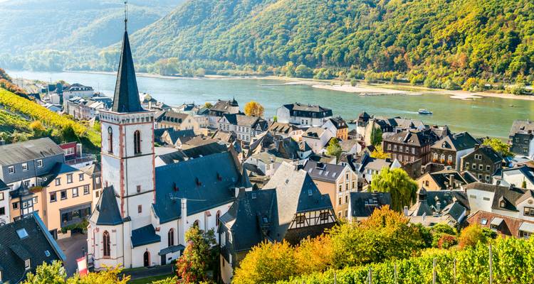 Village en bord de rivière avec un haut clocher d'église, des vignobles et de douces collines verdoyantes le long du Rhin.