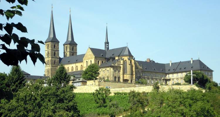 Abbaye perchée au sommet d'une colline avec deux flèches jumelées surplombant une verdure luxuriante sous un ciel bleu dégagé.