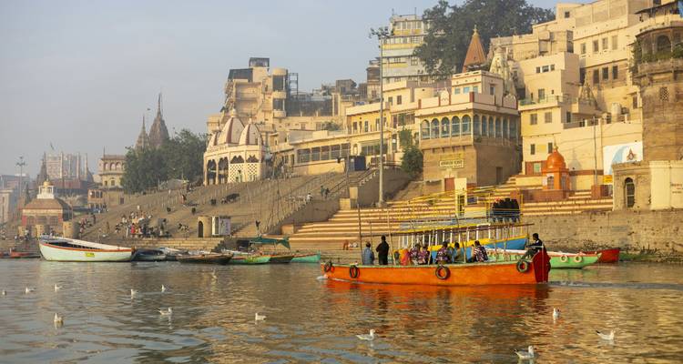 Colorful passenger boat gliding past the riverside ghats of an ancient city on the Ganges.