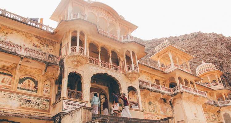 Visitors climb stone steps into an ornate, fresco-lined haveli set against rocky cliffs.