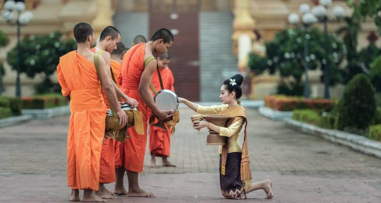 Moines recevant l'aumône d'une femme en tenue traditionnelle sur un chemin pavé.
