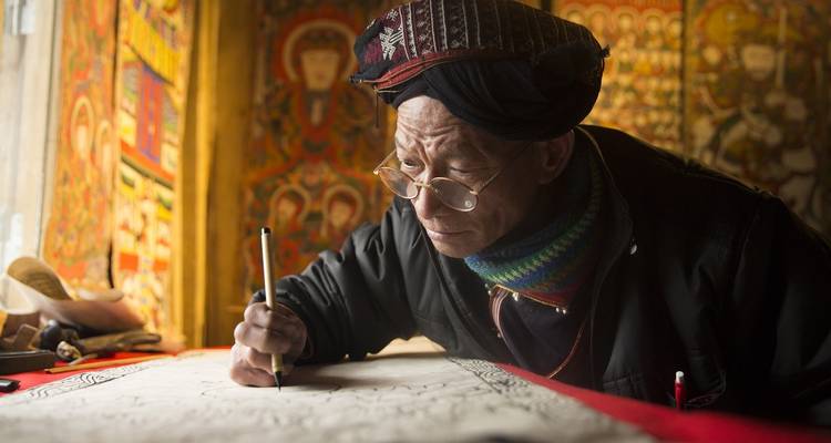 Un homme âgé dessinant avec attention en tenue traditionnelle dans une pièce colorée.