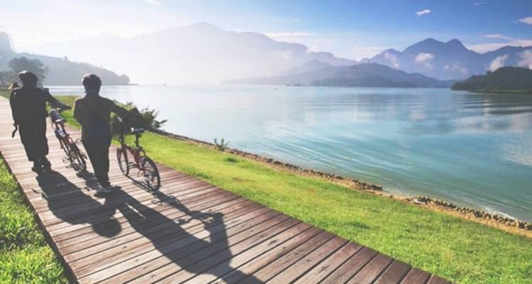 Two cyclists walk their bikes along a wooden path beside a tranquil lake with hazy mountains in back-light.