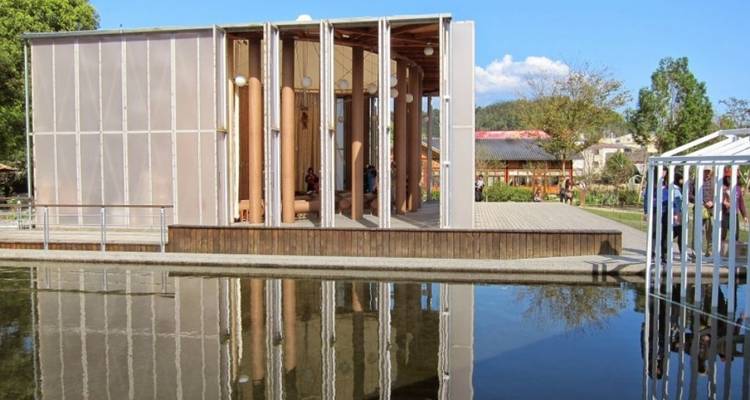 Contemporary pavilion with reflective water pool and blue skies, showing light wooden structure and visitors inside.